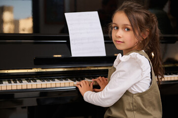 Cute child girl pianist sitting at grand piano, enjoys the rhythm of classical music while plays piano, smiles at camera