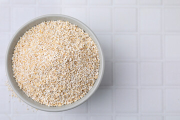 Dry barley groats in bowl on white tiled table, top view. Space for text