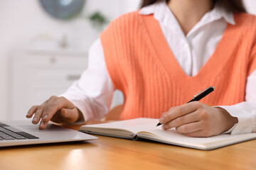 Young woman writing in notebook while working on laptop at wooden table, closeup