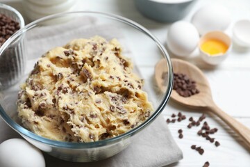 Chocolate chip cookie dough in bowl and ingredients on white wooden table, closeup
