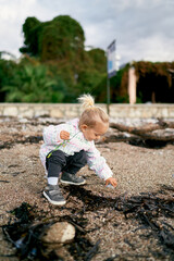 Little girl squats on the beach and touches a stone with her hand