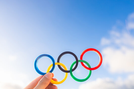 A hand holds iconic plastic Olympic rings against a vibrant blue sky, capturing the essence of the Olympic Games and International Olympic Committee. Ideal for those seeking content on the Olympics.