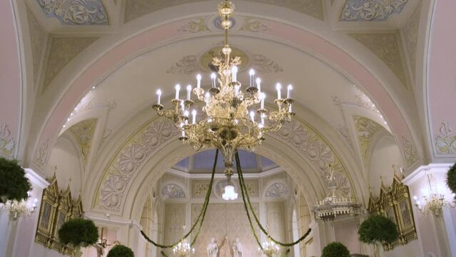 Enter Church On Wedding Ceremony Day. Chandeliers, Hanging Lights, Liturgical Decorations, Altar Adorned With Religious Symbols, Candles and Flowers. Painted Walls, Sculptures and Checkerboard Floor.