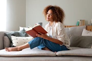 Pensive relaxed African american woman reading a book at home, drinking coffee sitting on the couch. Copy space.