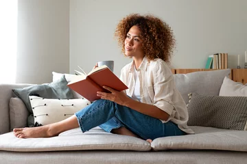 Foto op Canvas Pensive relaxed African american woman reading a book at home, drinking coffee sitting on the couch. Copy space. © Daniel