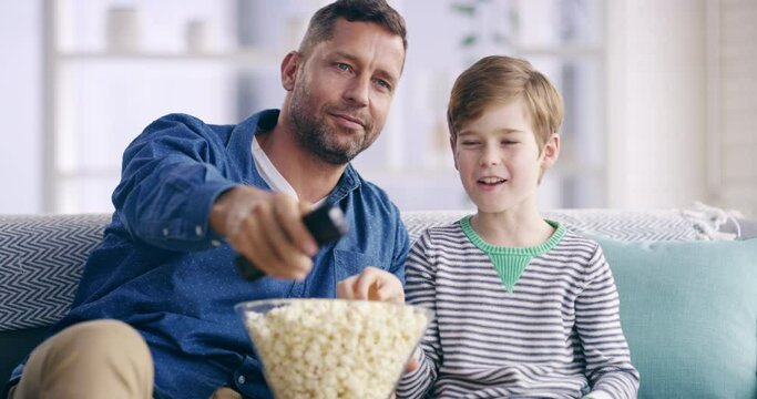 Family, popcorn and a man watching tv with his son on a sofa in the home living room together. Love, smile or happy with a father and boy child streaming a film, movie or video in an apartment