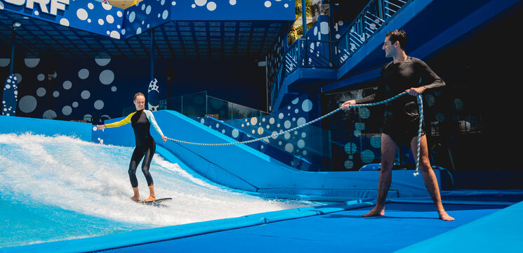 Beautiful young woman surfing with trainer on a wave simulator at a water amusement park