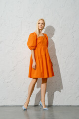 Fototapeta na wymiar Stylish adult woman in orange dress posing near white textured wall