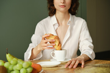 Unrecognisable woman dipping croissant into cappuccino in the morning at home