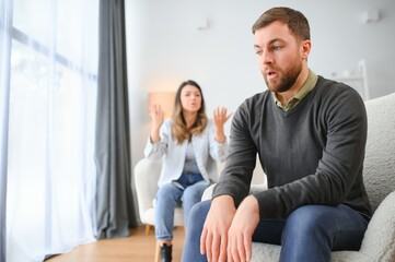 Couple arguing. Wife shouting to her desperate husband sitting on a couch in the living room at home