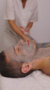 Portrait of a young man with cosmetic facial mask on his face, lying on couch while cosmetologist doing chest massage for him. Close-up, man on spa treatments. Vertical video.