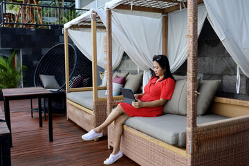 Fototapeta na wymiar happy beautiful asian woman in red dress sitting in gazebo rattan sofa chair outdoors while working with her laptop beside swimming pool