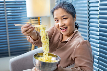 Senior Chinese woman using chopstick to eat noodle at home for cultural staple food and longevity...
