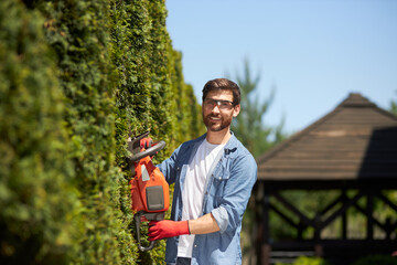 Professional gardener trimming, shaping evergreen thuja hedge with modern electric trimmer at summer time. Portrait of smiling worker looking at camera, while pruning. Concept of gardening services. - obrazy, fototapety, plakaty