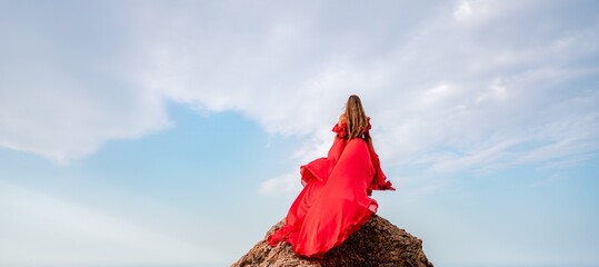 woman sky red dress. Woman with long hair on a sunny seashore in a red flowing dress, back view,...