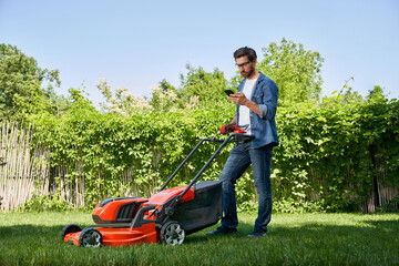 Focused male gardener in glasses using smartphone, while having break after trimming lawn in garden. Side view of serious guy with lawnmower working on backyard, against fence. Concept of gardening.