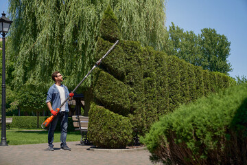 Strong male gardener in casual clothes trimming hedge with modern electric machine on backyard. Front view of caucasian man in gloves working with gardening tool outdoors. Concept of topiary. 
