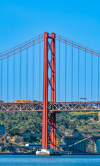 April 25 Bridge built of red steel like the Golden Gate, over the Tagus River in Lisbon, with car and truck traffic on a sunny day with clear blue sky. Portugal. Factories in the background.