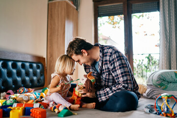 Dad and little girl are sitting on the bed touching their foreheads playing toys - obrazy, fototapety, plakaty