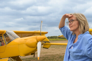 A blonde woman in glasses on the runway against the background of a vintage yellow plane and a stormy sky