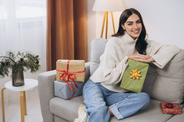 Beautiful happy woman unwrapping New Year or Christmas presents at home on sofa
