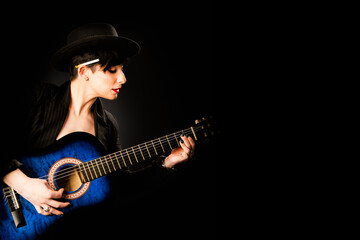 A young beautiful woman in black with a cigarette behind her ear plays an acoustic blue guitar