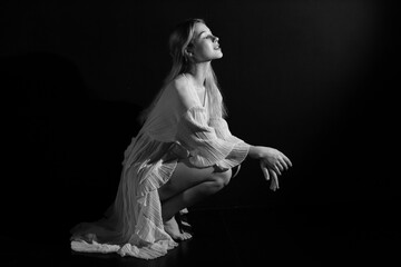 Fototapeta premium Low key black and white portrait of young pretty woman in vintage white dress sitting on the floor