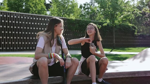 Two girls relax and drink fresh water from a plastic bottle after an active workout on a skateboard and scooter