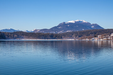 Naklejka premium Panoramic view on lake Faakersee in Austrian Alps, Carinthia, Austria. Lake is surrounded by high snow capped mountains. Calm water surface with reflections of landscape. Looking at Dobratsch peak