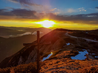 Summit cross with panoramic sunrise view from summit Dobratsch on Julian Alps and Karawanks in Austria, Europe. Silhouette of endless mountain ranges with orange colors of sky. Jagged sharp peaks