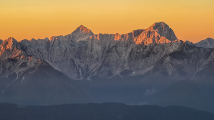 Panoramic sunrise view from summit Dobratsch on Julian Alps and Karawanks in Austria, Europe. Silhouette of endless mountain ranges with orange and pink colors of sky. Jagged sharp peaks and valleys - obrazy, fototapety, plakaty