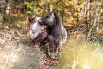 A young woman and her shetland pony cuddle together in a forest in autumn outdoors