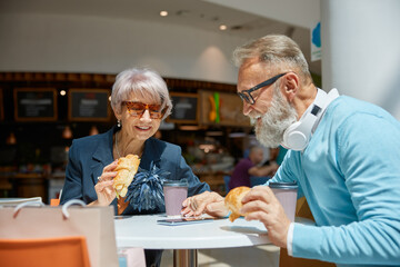 Happy retired couple eating croissants at cafeteria in shopping mall