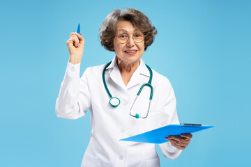 Portrait of gray haired senior woman, doctor holding diagnosis, writing recipe standing isolated on blue background. Concept of medicine, health care
