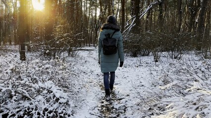 Young woman walking along path in sunny snowy forest. Girl with backpack going among trail at...