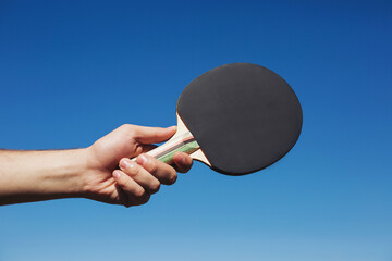 Hand holding black ping pong racket on blue sky background, closeup. Ping pong paddle. Playing ping...