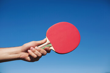 Hand holding red ping pong racket on blue sky background, closeup. Ping pong paddle. Playing ping...