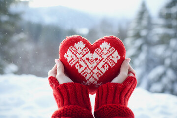 Female hands in red sweater holding knitted heart on snowy background.