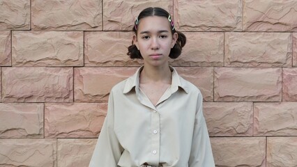 A teenage girl in a beige shirt stands near a peach-colored brick wall.