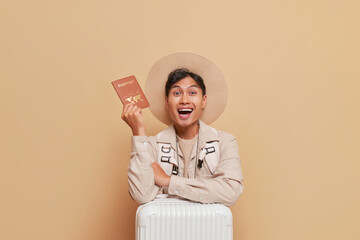 Happy chineese guy in broadbrim hat holds suitcase showing his passport smiling, journey time concept, copy space