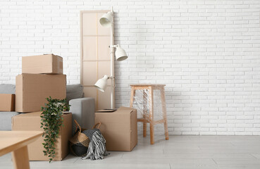 Cardboard boxes with plants and furniture near white brick wall in room on moving day
