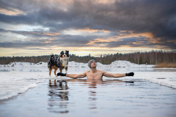 Young man taking an ice bath with his dog suring sunset. Swimming in a frozen lake in winter. Healthy cold swim. - obrazy, fototapety, plakaty