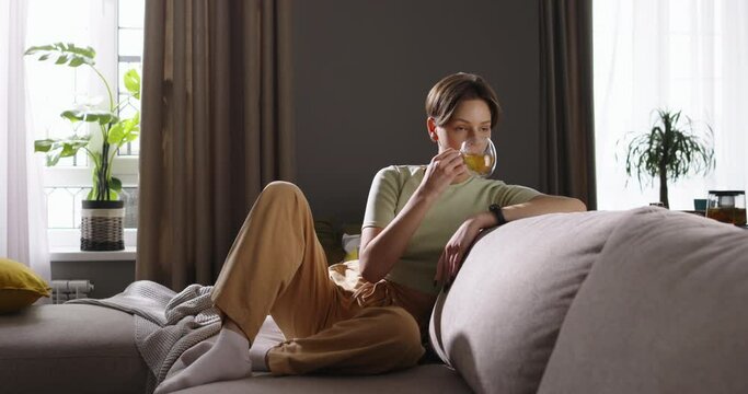 Happy female relaxed sitting on sofa in living room, basking in morning sun with mug in hand, smiling softly. Tranquility relaxation and serenity. Cinematic advertising. Cell gadget technology