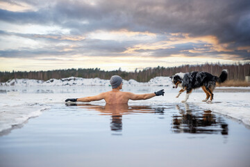 Young man taking an ice bath with his dog suring sunset. Swimming in a frozen lake in winter....