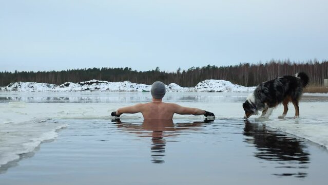 Young man taking an ice bath with his dog suring sunset. Swimming in a frozen lake in winter. Healthy cold swim.