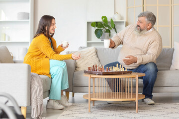 Mature man and nurse with cups of tea playing chess at home - obrazy, fototapety, plakaty