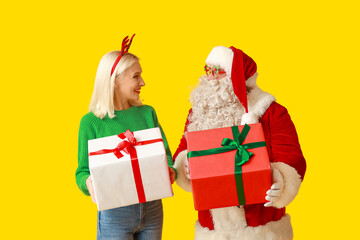 Santa Claus and beautiful mature woman in Christmas deer horns with gift boxes on yellow background