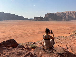 Young woman sitting recording with her mobile phone the sunset in the desert of Wadi Rum, Jordan....