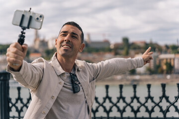 Young content creator filming himself with his cell phone on a tripod while talking to his followers. Boy filming with his cell phone pointing the river and the city of Budapest. - obrazy, fototapety, plakaty