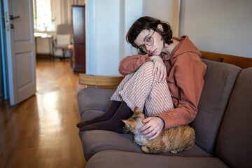 Lonely depressed teenager sitting on sofa, stroking beloved adorable breed cat Devon Rex. Teen girl in need of comfort, emotional support, empathy, contact. Fluffy pet performing antistress function
